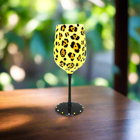 A wine glass with a yellow animal print design, displayed on a wooden surface with foliage in the background.