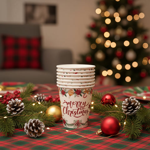 Stack of 'Merry Christmas' paper cups on a table with festive decorations and a blurred Christmas tree in the background.