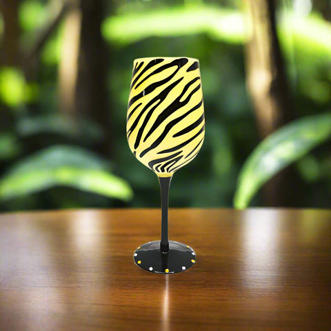 A wine glass with a yellow animal print design, displayed on a wooden surface with foliage in the background.